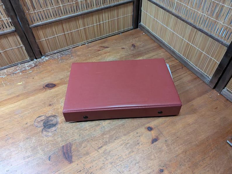 Op de afbeelding: Een rechthoekige, bordeauxrode doos met een gladde, leerachtige textuur. De doos heeft afgeronde hoeken en twee kleine, zilverkleurige klinknagels aan de voorkant. Hij staat op een houten oppervlak.