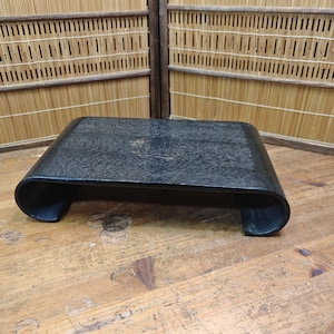 Puede incluir: Una mesa de centro rectangular negra con patas curvas. La mesa tiene una superficie texturizada y está hecha de un material oscuro. La mesa está colocada sobre una superficie de madera, con una pantalla de bambú en el fondo.