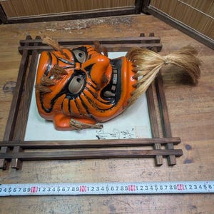 May include: An orange and black Japanese Noh mask with a fierce expression, mounted in a dark wooden frame. The mask has detailed features and a flowing, light-brown hairpiece. The frame rests on a wooden surface.