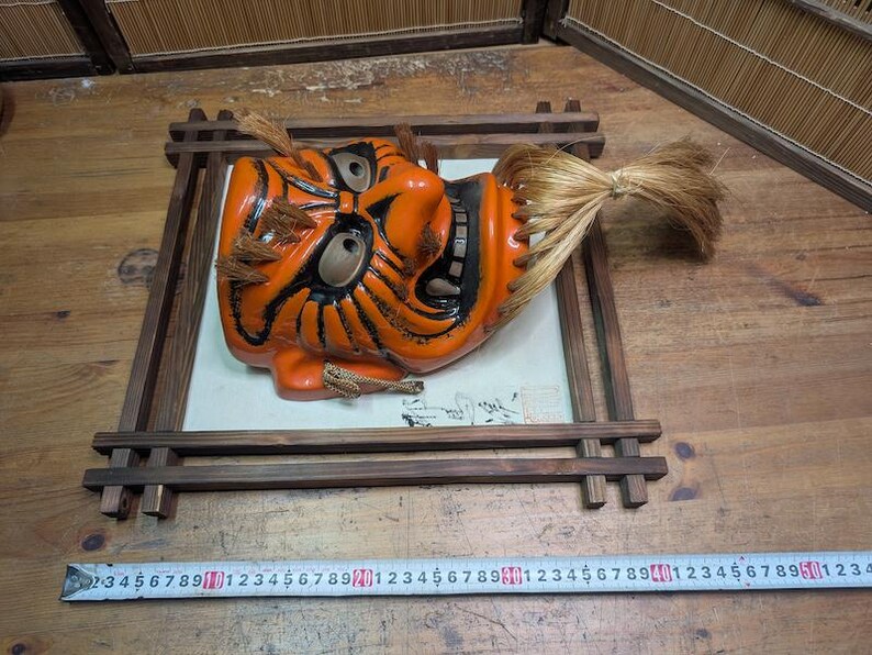 May include: An orange and black Japanese Noh mask with a fierce expression, mounted in a dark wooden frame. The mask has detailed features and a flowing, light-brown hairpiece. The frame rests on a wooden surface.