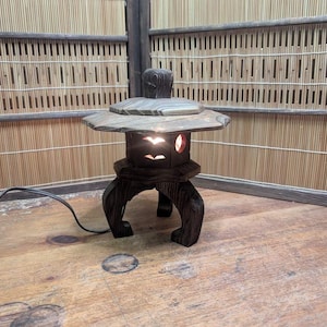 May include: A dark brown, wooden Japanese garden lantern with a stone-like top and carved bird-shaped cutouts. The lantern is lit from within, casting a warm glow. It has a three-legged base and a power cord.