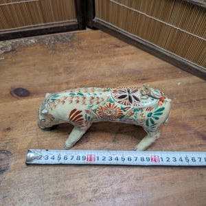 May include: A ceramic figurine of a boar, cream-colored with intricate floral and geometric patterns in red, green, and brown. The boar is approximately 4 inches long, with a ruler in the foreground.