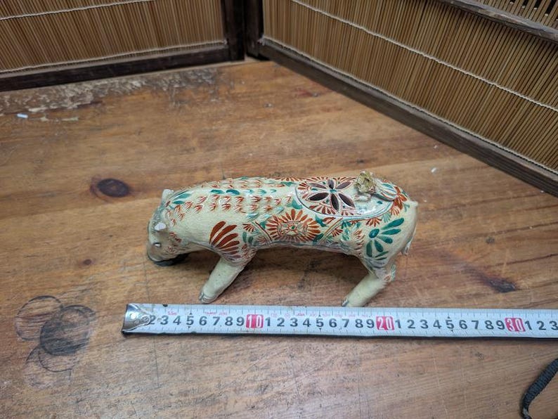 May include: A ceramic figurine of a boar, cream-colored with intricate floral and geometric patterns in red, green, and brown. The boar is approximately 4 inches long, with a ruler in the foreground.