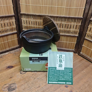 May include: A black cast iron cooking pot with a lid, on a green box. A card with Japanese text is in front of the pot. The pot has a handle and is designed for cooking.