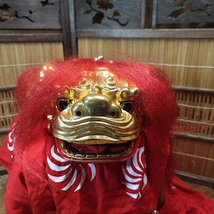 以下が含まれることがあります： 金色の顔と赤い毛並みが特徴的な、日本の伝統的な獅子舞のマスク。木製のパネルと竹製のブラインドを背景に、白いアクセントが施された赤い衣装を着ています。