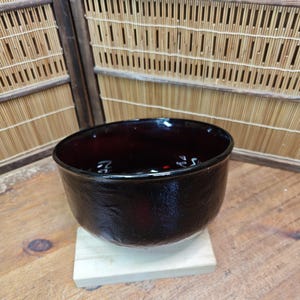 Könnte beinhalten: Eine dunkelburgunderfarbene Schale mit strukturierter Oberfläche, die auf einem kleinen, hellen Holzblock steht. Die Schale hat eine breite, offene Oberseite und eine abgerundete Form. Der Hintergrund zeigt einen Bambus-Bildschirm.