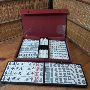 Op de afbeelding: Een rode Mahjong-set met witte tegels met zwarte en rode symbolen. De set wordt open gepresenteerd en toont geordende rijen tegels in een rode koffer. Extra tegels zijn gerangschikt in een apart zwart dienblad.