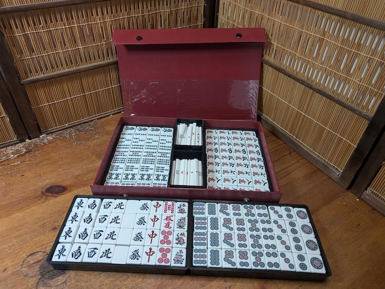 Op de afbeelding: Een rode Mahjong-set met witte tegels met zwarte en rode symbolen. De set wordt open gepresenteerd en toont geordende rijen tegels in een rode koffer. Extra tegels zijn gerangschikt in een apart zwart dienblad.