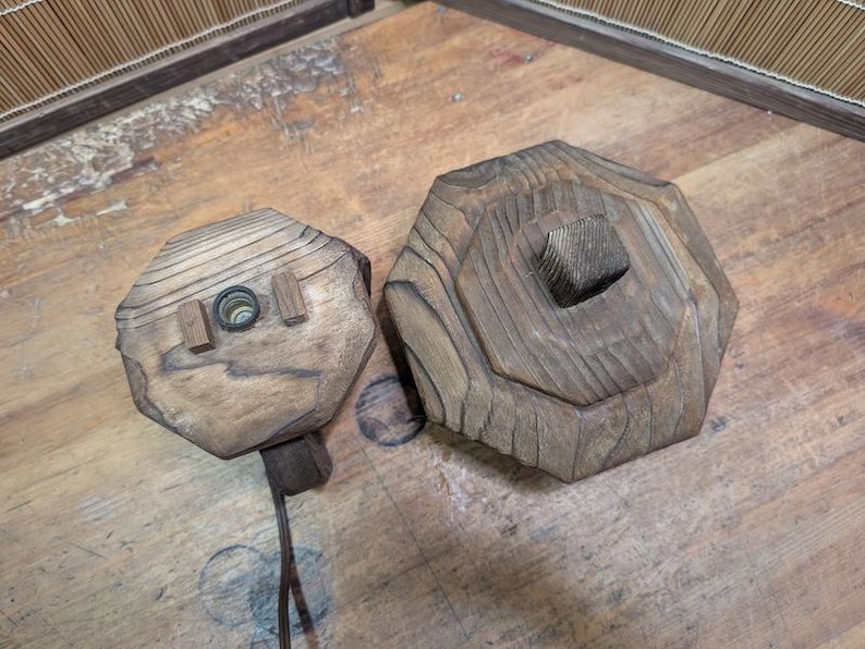 May include: Two wooden, hexagonal-shaped objects. One has a hole with a light fixture and two small wooden blocks. The other has a square hole in the center. Both are made of light brown wood with visible grain.
