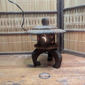 May include: A dark brown wooden Japanese lantern with a curved top and a base with three curved legs. The lantern has a crescent moon and bird cutouts, and a light shines from within. The lantern is on a wooden surface.