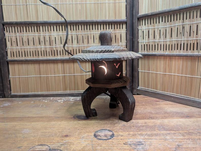 May include: A dark brown wooden Japanese lantern with a curved top and a base with three curved legs. The lantern has a crescent moon and bird cutouts, and a light shines from within. The lantern is on a wooden surface.