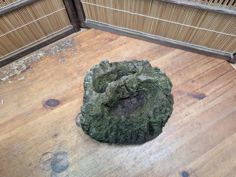 Puede incluir: Roca texturizada, cubierta de musgo verde, con una depresi&oacute;n central. Los colores dominantes son el gris y el verde, lo que sugiere un aspecto natural y envejecido. Podr&iacute;a ser un elemento decorativo.