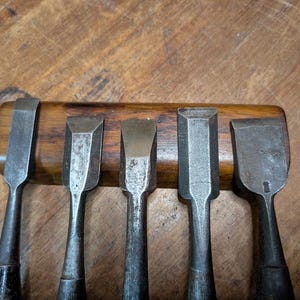 Puede incluir: Un juego de cinco cinceles de madera antiguos con hojas de metal y mangos de madera oscura. Los cinceles est&aacute;n dispuestos en fila, con las hojas hacia arriba. El fondo es una superficie de madera.