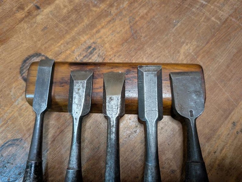 Puede incluir: Un juego de cinco cinceles de madera antiguos con hojas de metal y mangos de madera oscura. Los cinceles est&aacute;n dispuestos en fila, con las hojas hacia arriba. El fondo es una superficie de madera.