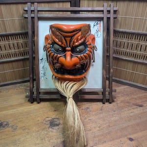 May include: An orange and black Japanese Noh mask with a long, light-colored fiber beard, framed in a dark wooden structure. The mask has detailed features and is mounted on a white background with Japanese calligraphy.