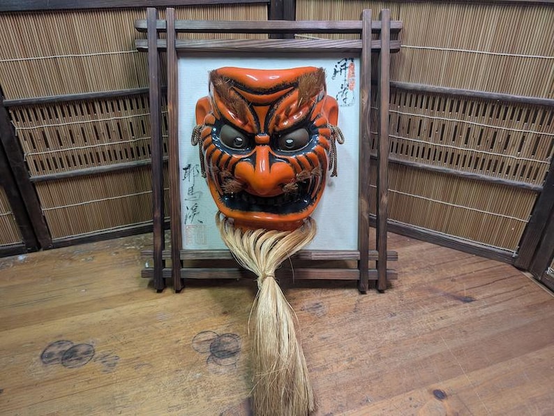 May include: An orange and black Japanese Noh mask with a long, light-colored fiber beard, framed in a dark wooden structure. The mask has detailed features and is mounted on a white background with Japanese calligraphy.