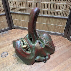 Op de afbeelding: Een gedetailleerd, handgeschilderd masker met een uniek ontwerp. Het masker heeft een prominente, omhoog gebogen aanhangsel en is geschilderd in tinten rood, groen en goud. Het masker ligt op een houten oppervlak.