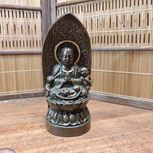 May include: Bronze-colored statue of a seated figure with hands clasped in prayer, surrounded by smaller figures. The statue has a halo and sits atop a lotus flower base. The background features a bamboo screen and wooden surface.