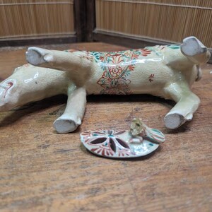 May include: A ceramic figurine of a pig, lying on its side, with a separate, decorative, oval-shaped piece. The pig is cream-colored with red, green, and blue floral patterns. The oval piece has a similar design.