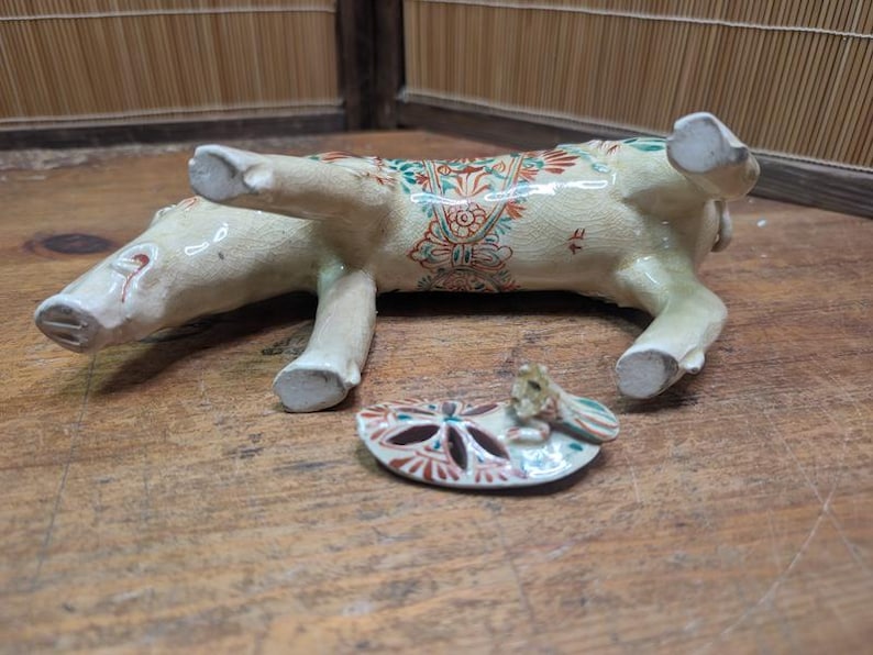 May include: A ceramic figurine of a pig, lying on its side, with a separate, decorative, oval-shaped piece. The pig is cream-colored with red, green, and blue floral patterns. The oval piece has a similar design.