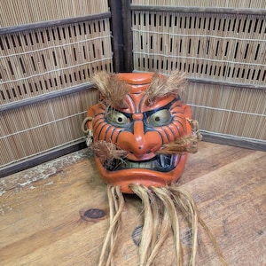 Könnte beinhalten: Eine orangefarbene japanische Dämonenmaske mit goldenen Augen, schwarzen Markierungen und braunen Haar-Akzenten. Die Maske hat einen grimmigen Ausdruck und wird auf einer Holzoberfläche präsentiert. Die Maske ist eine traditionelle japanische Kunstform.