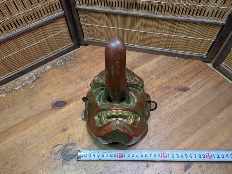 Op de afbeelding: Een vintage Japanse Tengu-masker, met een lange neus en een felle uitdrukking. Het masker is geschilderd in groen, rood en goud, met tekenen van slijtage. Het is een traditioneel item dat wordt gebruikt in de Japanse folklore en theater.