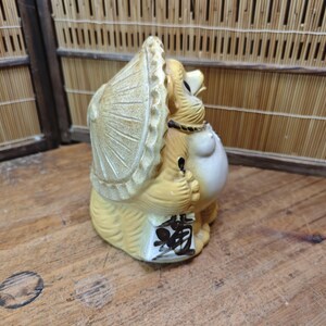 May include: Tanuki statue, a traditional Japanese raccoon dog figurine. The ceramic figure is tan and cream-colored, wearing a hat and holding a sake bottle. The base has Japanese characters. A charming decorative item.