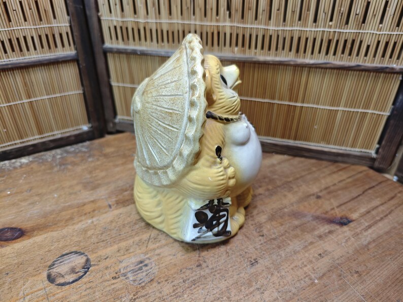 May include: Tanuki statue, a traditional Japanese raccoon dog figurine. The ceramic figure is tan and cream-colored, wearing a hat and holding a sake bottle. The base has Japanese characters. A charming decorative item.