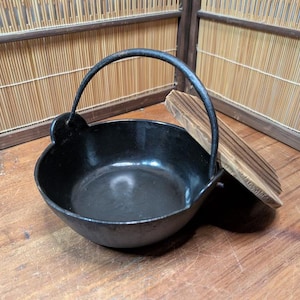 Könnte beinhalten: Ein schwarzer gusseiserner Topf mit gebogenem Griff und Holzdeckel. Der Topf hat eine runde Form und steht auf einer Holzoberfläche. Der Deckel besteht aus hellem Holz mit dunklen Streifen.