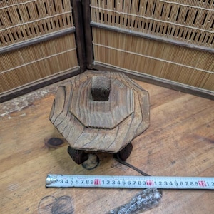 May include: A weathered, wooden decorative object with a geometric design. The object is brown and has a square top with a series of concentric rings. It sits on a small, dark base. A metal measuring tape is nearby.