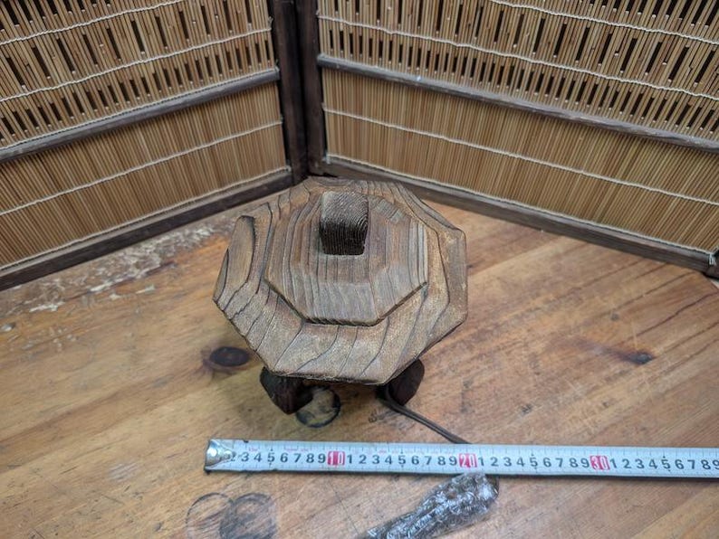 May include: A weathered, wooden decorative object with a geometric design. The object is brown and has a square top with a series of concentric rings. It sits on a small, dark base. A metal measuring tape is nearby.