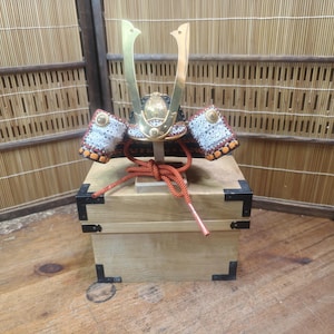 May include: A Japanese samurai helmet, or kabuto, with gold-coloured horns and intricate detailing. The helmet rests on a wooden box with black corner reinforcements. A red cord is tied around the base of the helmet.