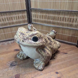 Könnte beinhalten: Keramikfrosch-Skulptur mit einem großen Frosch und zwei kleineren Fröschen auf dem Rücken. Der Frosch ist beige mit braunen Mustern und schwarzen Augen. Die Skulptur steht auf einer Holzoberfläche.