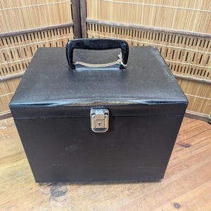 K&ouml;nnte beinhalten: Eine schwarze, quadratische Aufbewahrungsbox mit Griff und Metallverschluss. Das Geh&auml;use hat eine strukturierte Oberfl&auml;che und einen schwarzen Griff. Der Verschluss ist silberfarben. Das Geh&auml;use steht auf einer Holzoberfl&auml;che.