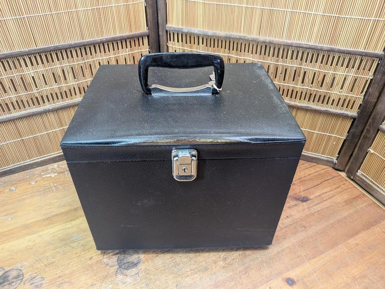 K&ouml;nnte beinhalten: Eine schwarze, quadratische Aufbewahrungsbox mit Griff und Metallverschluss. Das Geh&auml;use hat eine strukturierte Oberfl&auml;che und einen schwarzen Griff. Der Verschluss ist silberfarben. Das Geh&auml;use steht auf einer Holzoberfl&auml;che.