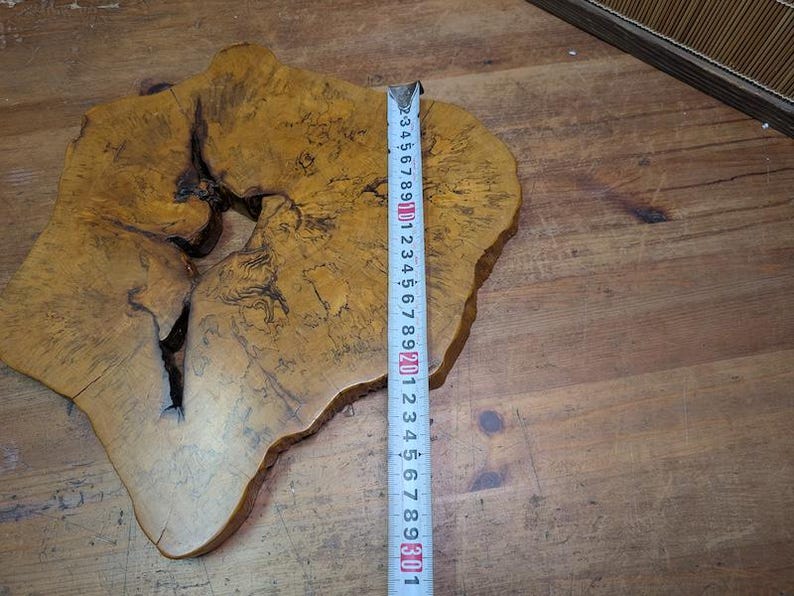 May include: A natural wood slab with a unique, irregular shape. The slab has a light brown color with darker grain patterns and a large, dark knot. A ruler shows the slab is approximately 12 inches wide.