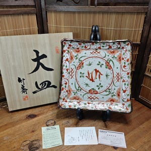 May include: A square, decorative ceramic plate with a floral and fish design in red, green, and white. The plate is displayed on a black stand, next to a wooden box with Japanese calligraphy.