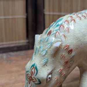 May include: A ceramic figurine of an animal, possibly a bear or dog, with a cream-colored body and intricate floral and leaf patterns in red, teal, and green. The figurine has a cracked glaze finish.