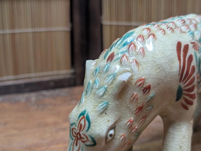 May include: A ceramic figurine of an animal, possibly a bear or dog, with a cream-colored body and intricate floral and leaf patterns in red, teal, and green. The figurine has a cracked glaze finish.