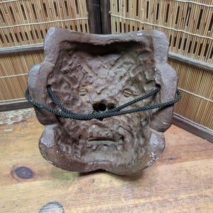 Op de afbeelding: Een verweerde, bruine masker met een getextureerd oppervlak en een donkergroen koord. Het masker heeft een gestileerd gezicht met een kleine mond en ooggaten. Het masker is waarschijnlijk gemaakt van een hard materiaal.