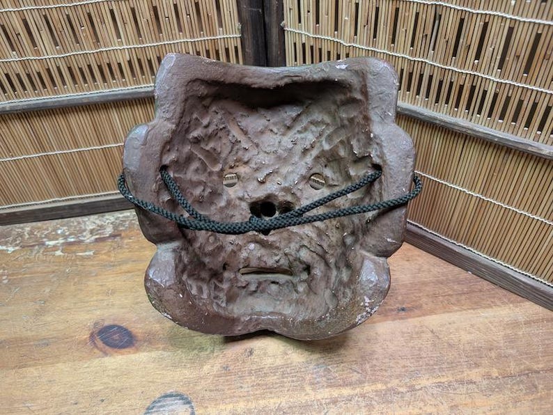 Op de afbeelding: Een verweerde, bruine masker met een getextureerd oppervlak en een donkergroen koord. Het masker heeft een gestileerd gezicht met een kleine mond en ooggaten. Het masker is waarschijnlijk gemaakt van een hard materiaal.