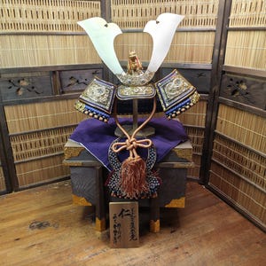 May include: A traditional Japanese samurai helmet, with a silver crest and intricate detailing. The helmet is displayed on a wooden stand with a purple cloth, and a small wooden plaque with Japanese characters. The background includes bamboo screens.