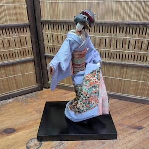 May include: A Japanese geisha doll in a light blue kimono with a colorful floral pattern on the hem. The doll has a black hairstyle with decorative hairpins and is displayed on a black square base.