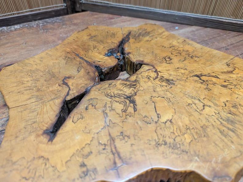 May include: Close-up of a unique, irregularly shaped wooden slab. The light brown surface features dark, intricate grain patterns and a deep, dark fissure running through the center. The wood's natural edges are visible.