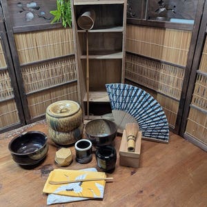 May include: A collection of tea ceremony items including a lidded ceramic jar, two dark glazed bowls, a small black cup, a bamboo whisk in a wooden box, a folding fan with a blue geometric pattern, and a yellow cloth with a crane illustration.