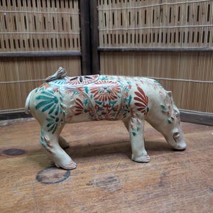 May include: A ceramic sculpture of a pig with a lid, featuring a cream-colored base with intricate floral patterns in teal and red. The pig is in a walking pose, with its head lowered. The sculpture is on a wooden surface.