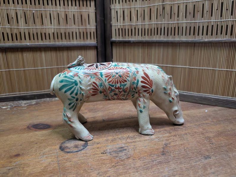 May include: A ceramic sculpture of a pig with a lid, featuring a cream-colored base with intricate floral patterns in teal and red. The pig is in a walking pose, with its head lowered. The sculpture is on a wooden surface.