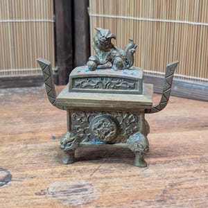 Puede incluir: Un quemador de incienso de bronce detallado con una figura decorativa de león en la parte superior. El quemador presenta tallas ornamentadas, patas con cabeza de león y asas con diseño geométrico. Se exhibe sobre una superficie de madera.