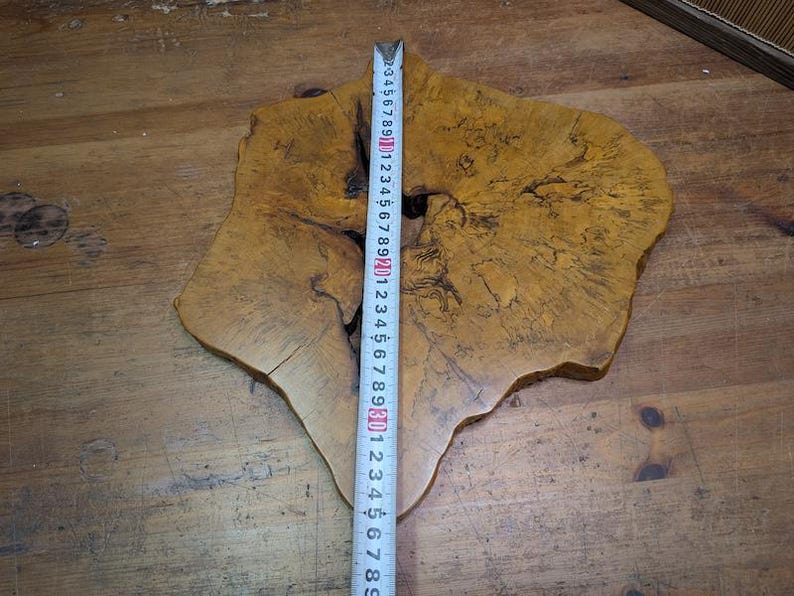 May include: A uniquely shaped, natural wood slab with a light brown color and dark grain patterns. A measuring tape shows the slab is approximately 30 inches long. The wood has a rough, organic edge and a hole in the center.