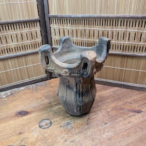 May include: A weathered, hand-carved, ceramic vessel with a unique, open-top design. The vessel features a flared rim with four rounded protrusions and intricate carved patterns on the body. The color is a mix of brown and gray.